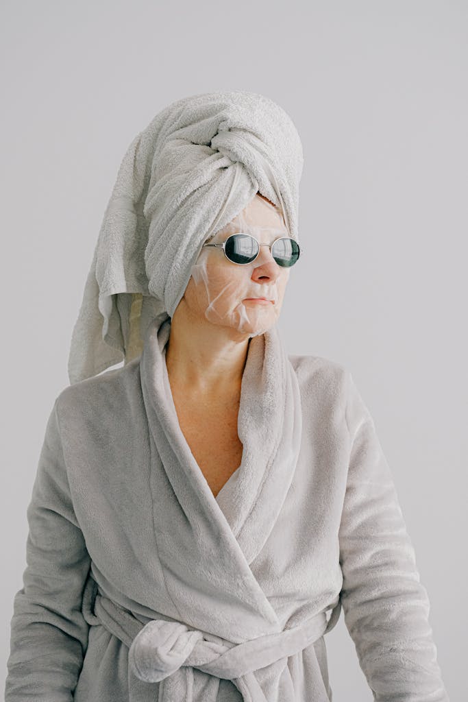 Aged female in gray bathrobe and with towel on head standing on gray background and looking away in sunglasses with sheet mask on face