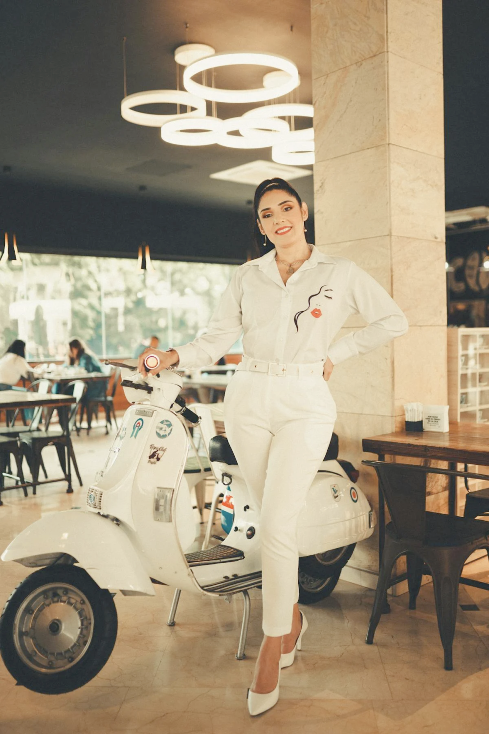 a woman wearing white pant shirt posing with a scooter