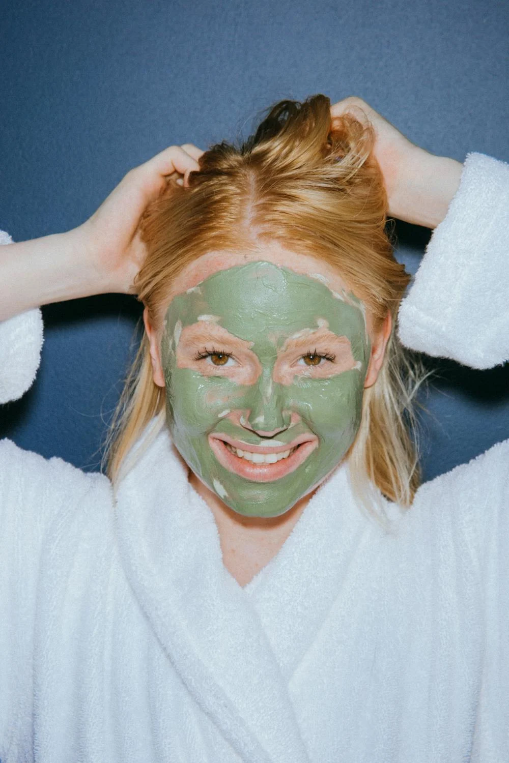 closeup of a smiling woman with a green face pack on, wearing a bathrobe