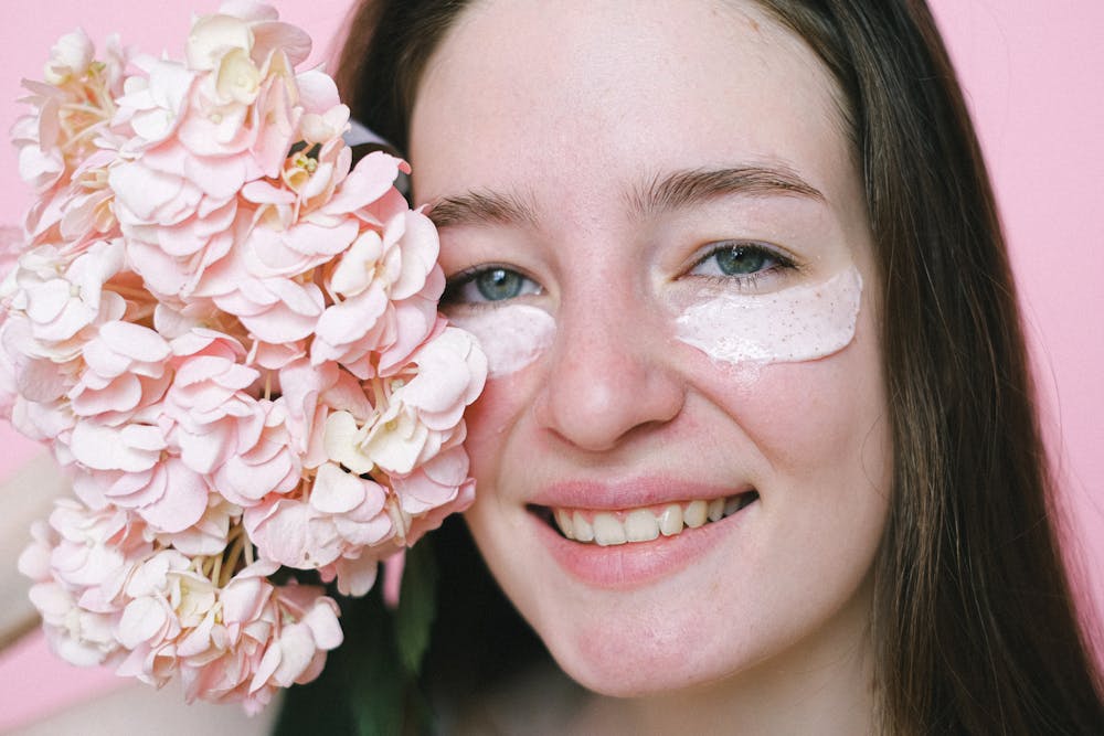 Crop positive young female model with eyes mask smiling at camera while demonstrating hydrangea flower on pink background